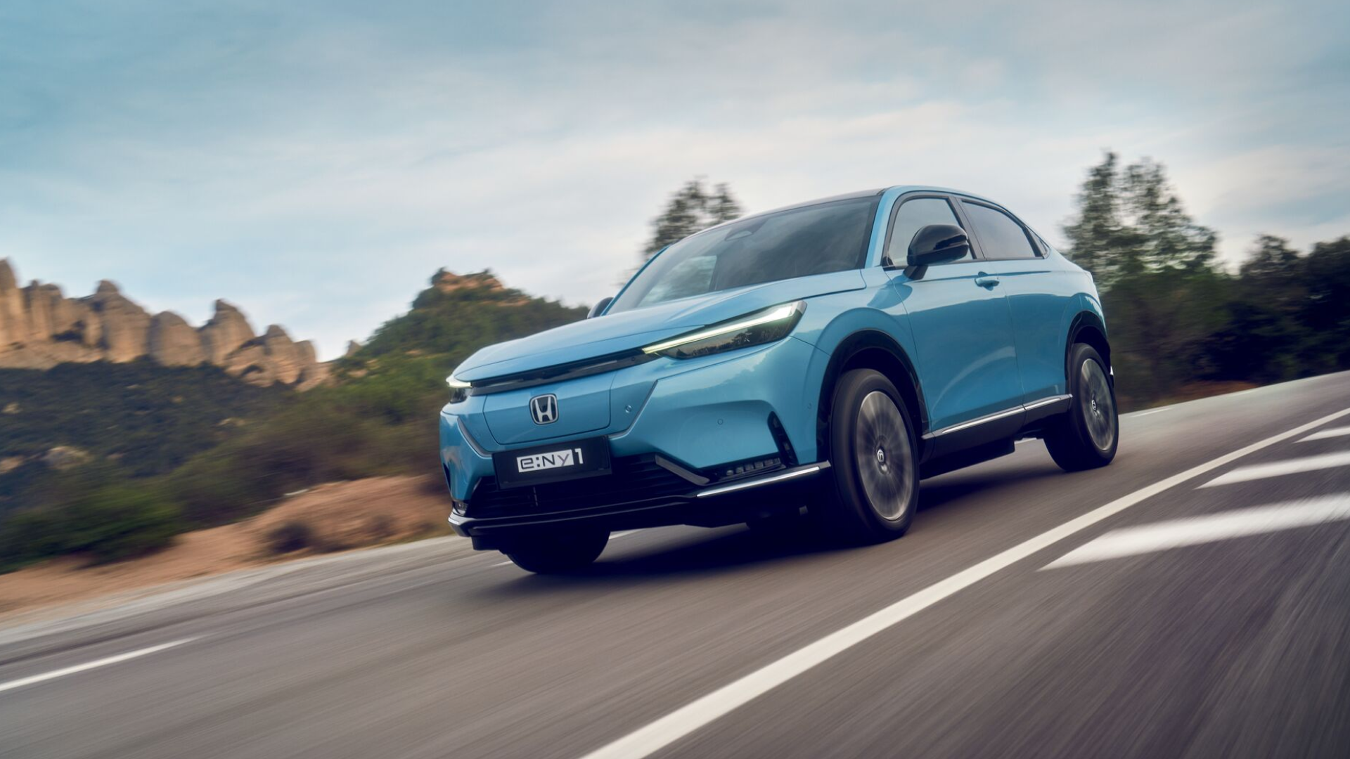 A light blue Honda e:NY1 electric SUV driving on a winding road with rocky terrain and trees in the background. The vehicle's sleek design and illuminated headlights are prominently featured as it moves along the road.